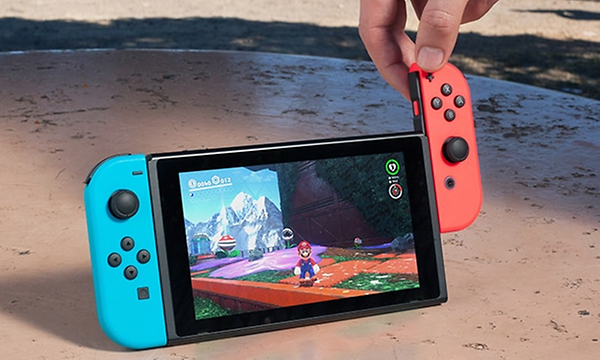 Nintendo Switch på stativ på ett bord och super mario på skärmen. En hand är på väg att ta bort den ena joycon-kontrollen. 